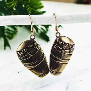 May include: A pair of gold-tone earrings with a unique design featuring a textured, oval-shaped pendant with a geometric pattern. The earrings have a simple hook closure.