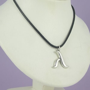 Puede incluir: Un collar de plata con un colgante de la letra 'A' estilizada en un cordón de cuero negro.