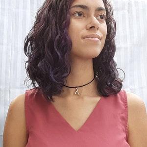 Op de afbeelding: Een persoon met krullend paars haar draagt een rood, mouwloos topje en een zwarte choker met een zilveren hanger.