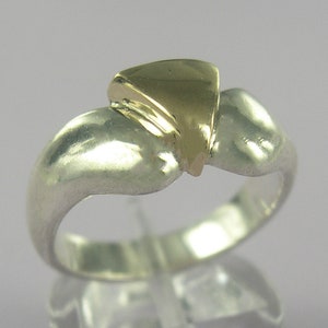 May include: A silver ring with a gold-colored, triangular accent. The ring has a smooth, polished finish and a unique, abstract design. The gold accent is raised and adds a contrasting element to the silver band.