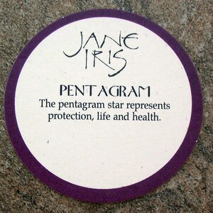 May include: A white coaster with a purple border and the text "JANE IRIS PENTAGRAM The pentagram star represents protection, life and health."