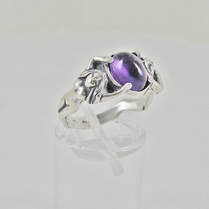 Peut inclure: Une bague en argent ornée d'une grosse pierre ovale violette. La bague présente des figures sculptées de chaque côté de la pierre. La bague est présentée sur un support transparent sur fond blanc.