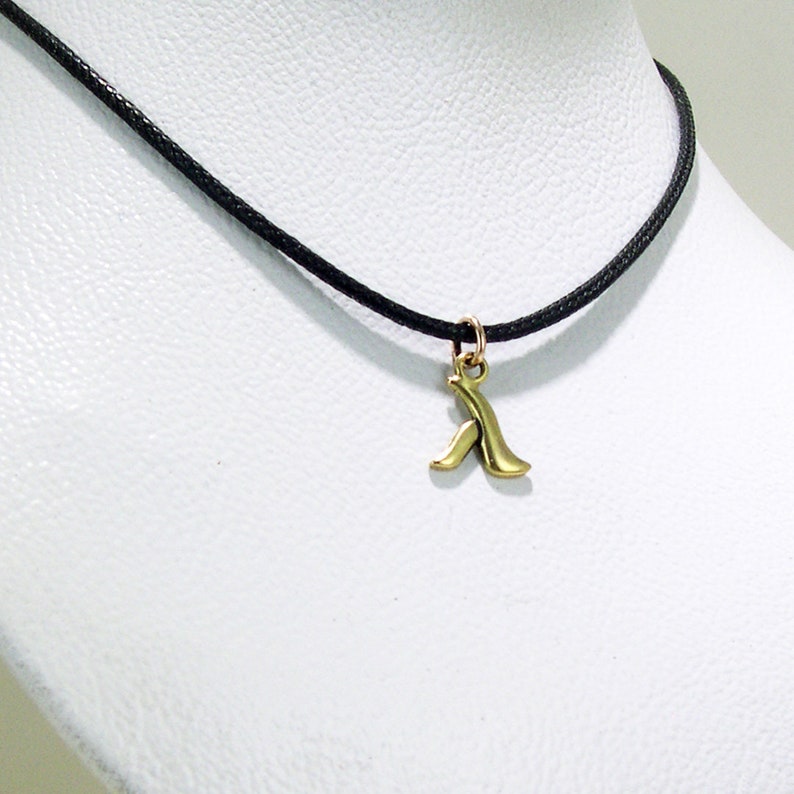 K&ouml;nnte beinhalten: Eine schwarze Schnurkette mit einem goldenen Anh&auml;nger in Form eines stilisierten Buchstabens A.