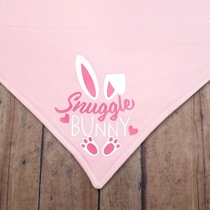 May include: A light pink pet bandana with the text "Snuggle Bunny" and bunny ears, hearts, and paw prints. The bandana is triangular and made of fabric. The text "SYCBSIRIUS" is printed on the top right corner.