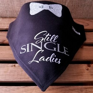 May include: Black fabric bib with a silver bow and the text "Still Single Ladies" in silver glitter.
