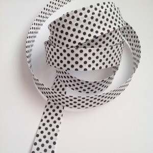 Op de afbeelding: Een wit lint met een herhalend patroon van zwarte stippen is opgerold op een witte ondergrond. Het lint is gemaakt van een lichte stof en is ideaal voor knutselen, cadeauverpakking of om een decoratief tintje toe te voegen. Het lint is ongeveer 2,5 cm breed.