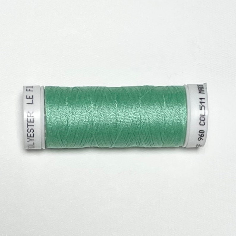Puede incluir: Una bobina de hilo de poli&eacute;ster verde claro. La bobina tiene etiquetas blancas con texto negro que dice "POLYESTER LE FIL" y "960 COL 511 MADE". El hilo est&aacute; bien enrollado y parece liso.