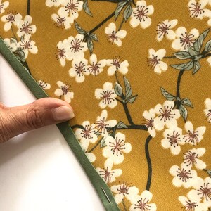 May include: Close-up of a fabric with a floral pattern. The fabric has a mustard yellow background with white cherry blossoms and green leaves. A green bias tape is sewn along the edge, held in place with pins.