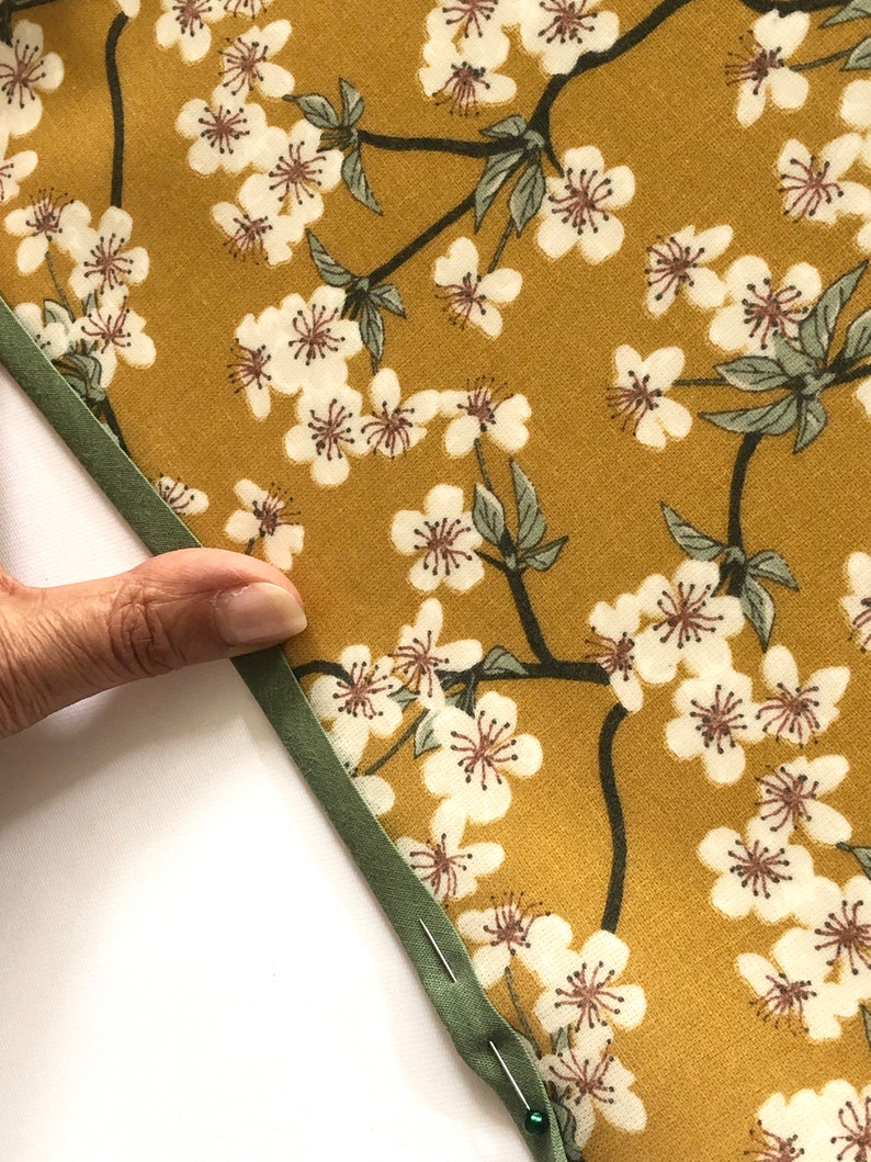 May include: Close-up of a fabric with a floral pattern. The fabric has a mustard yellow background with white cherry blossoms and green leaves. A green bias tape is sewn along the edge, held in place with pins.