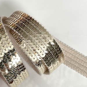 Peut inclure: Gros plan d'un ruban décoratif orné de paillettes dorées. Les paillettes circulaires sont densément disposées, créant un effet scintillant. Le ruban a un dos beige et est enroulé, révélant sa texture et son design.