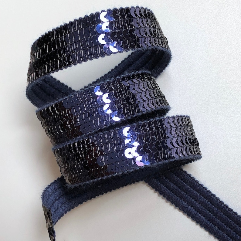 May include: Navy blue sequin ribbon, coiled on a white surface. The ribbon is made of small, shiny, dark blue sequins. The ribbon is approximately 1 inch wide. The ribbon is suitable for crafts, sewing, and embellishments.