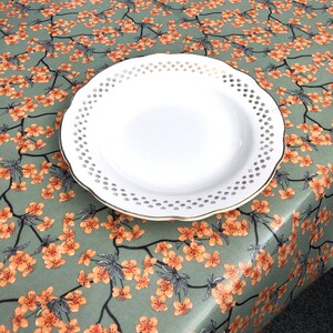 May include: A white ceramic plate with a gold rim and a decorative floral pattern. The plate is set on a tablecloth with a repeating pattern of orange blossoms and green leaves.