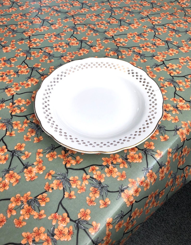 May include: A white ceramic plate with a gold rim and a decorative floral pattern. The plate is set on a tablecloth with a repeating pattern of orange blossoms and green leaves.