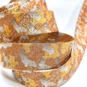 Puede incluir: Primer plano de una cinta decorativa con brillo metálico. La cinta presenta un patrón de colores oro, plata y bronce. La cinta está enrollada, mostrando su textura y diseño. Ideal para manualidades y envolver regalos.