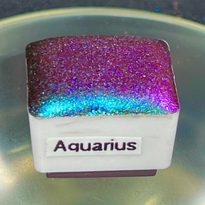May include: A small square container with a purple and blue iridescent glitter top. The container is labeled "Aquarius".