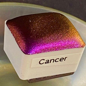 Puede incluir: Una pastilla de acuarela cuadrada con una etiqueta que dice "Cancer". La pastilla tiene un acabado brillante e iridiscente que cambia de dorado a rosa.