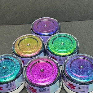 Pu&ograve; includere: Sei piccoli vasetti trasparenti pieni di glitter iridescenti in vari colori, tra cui viola, verde e verde acqua. Ogni vasetto ha un coperchio trasparente e un'etichetta bianca con testo e un motivo floreale. I vasetti sono disposti su una superficie scura.