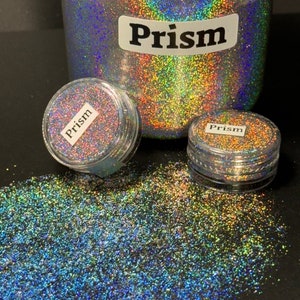 May include: A close-up of iridescent glitter in a jar labeled "Prism". The glitter is a mix of blue, green, and gold colors. There are two smaller jars of the same glitter in the foreground.