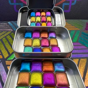 Puede incluir: Un juego de 16 acuarelas iridiscentes en una lata de metal. Las pinturas están dispuestas en una cuadrícula de 4x4 y tienen una variedad de colores, incluyendo azul, verde, morado y dorado. La lata tiene una etiqueta que dice "EmberLily Artisan Watercolors".