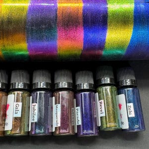 May include: Eight small bottles of iridescent glitter, labeled Gala, Jubilee, Masquerade, Parade, Revel, Zulu, and two others. The bottles are arranged in a row on a dark surface.