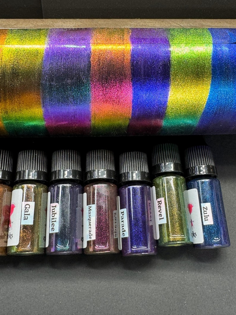 May include: Eight small bottles of iridescent glitter, labeled Gala, Jubilee, Masquerade, Parade, Revel, Zulu, and two others. The bottles are arranged in a row on a dark surface.