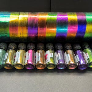 Könnte beinhalten: Zwölf kleine Glasflaschen mit irisierendem Lack, jede mit einer anderen Farbe und einem Namensschild. Die Flaschen sind in einer Reihe auf einer grauen Oberfläche angeordnet. Die Namen auf den Flaschen sind: "Fluorescent", "Festival", "Far Tuesday", "Jubilee", "Isomeral", "Parade", "Pearl", "Gala", "Flamboyant", "Gobi", "Labyrinth" und "Fluorescent".