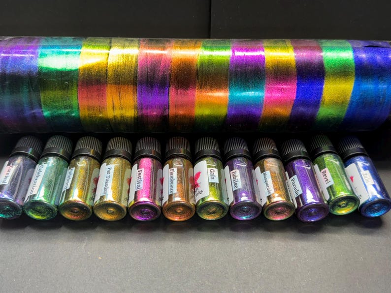 May include: Twelve small glass bottles with iridescent paint, each with a different color and name label. The bottles are lined up in a row on a gray surface. The names on the bottles are: "Fluorescent", "Festival", "Far Tuesday", "Jubilee", "Isomeral", "Parade", "Pearl", "Gala", "Flamboyant", "Gobi", "Labyrinth", and "Fluorescent".