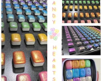 May include: A set of watercolor paints in small square pans, arranged in rows on a black surface. The pans are filled with shimmering, iridescent colors, including pink, blue, green, and gold. The text "CANDY HEARTS" is visible in the image.