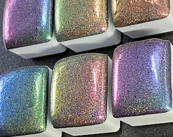 May include: Six square watercolor paint pans in a white holder. The paints have a shimmering, iridescent finish in shades of purple, green, gold, and copper. The paints are arranged in two rows of three.