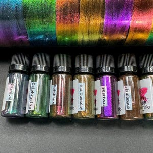 May include: Eight small glass bottles with black caps and labels. The labels have names like "Carnival", "Celebration", "Epiphany", "Fat Tuesday", "Festival", "Flambeaux", and "Shift Inks". The bottles contain iridescent glitter.