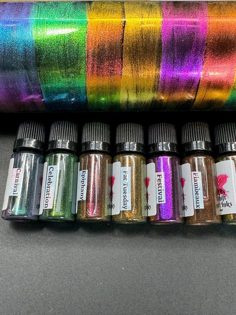 May include: Eight small glass bottles with black caps and labels. The labels have names like "Carnival", "Celebration", "Epiphany", "Fat Tuesday", "Festival", "Flambeaux", and "Shift Inks". The bottles contain iridescent glitter.