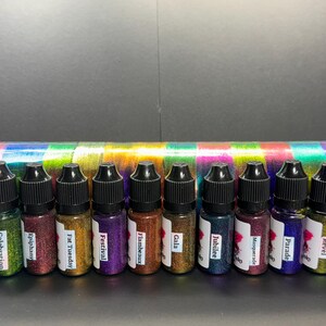 May include: A row of 12 small bottles filled with colorful glitter. The bottles are labeled with names like "Carnival", "Celebration", "Epiphany", "Fat Tuesday", "Festival", "Flambeaux", "Gala", "Jubilee", "Masquerade", "Parade", "Revel", and "Zulu".