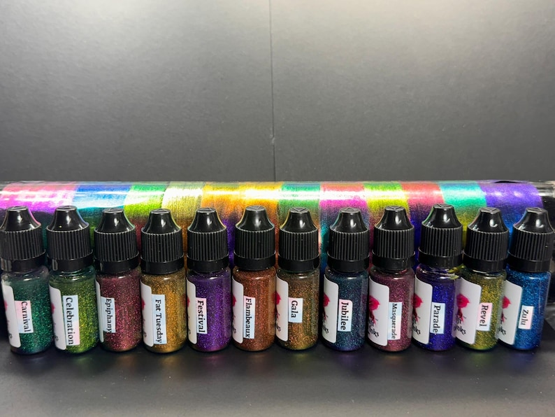 May include: A row of 12 small bottles filled with colorful glitter. The bottles are labeled with names like "Carnival", "Celebration", "Epiphany", "Fat Tuesday", "Festival", "Flambeaux", "Gala", "Jubilee", "Masquerade", "Parade", "Revel", and "Zulu".