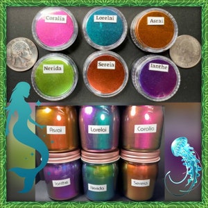 May include: Six small jars of iridescent pigment powder, labeled with names like "Coralia", "Lorelai", "Asrai", "Nerida", "Sereia", and "Ianthe". The jars are arranged in two rows of three, with a mermaid illustration and a jellyfish illustration in the background.