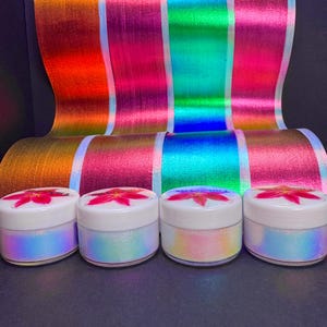 May include: Four small jars with white lids and iridescent glitter are in front of a colourful, metallic backdrop. Each jar has a red flower design on the lid. The backdrop has vertical stripes of orange, pink, green, and blue.