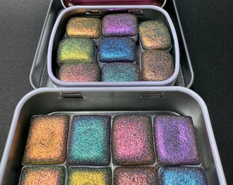 Puede incluir: Tres latas metálicas que contienen pastillas de pintura de acuarela iridiscente. La lata superior es rosa, la lata del medio es lavanda y la lata inferior es plateada. Cada lata contiene 12 pastillas de pintura.