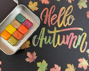 May include: A metal tin filled with nine watercolor paint pans. The pans are arranged in a grid pattern and are filled with various shades of orange, yellow, and green. The tin is open and the paint pans are visible. The background is black with the words "Hello Autumn" written in gold glitter.