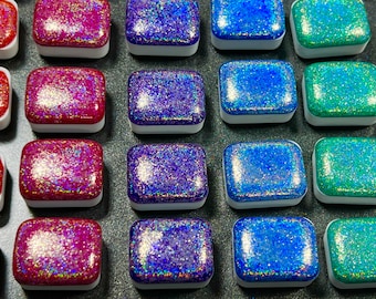 May include: A collection of 15 small square containers filled with iridescent glitter. The containers are arranged in three rows of five, with each row featuring a different color of glitter: pink, blue, and green.
