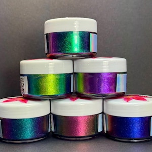 May include: A stack of six small jars with iridescent, color-shifting pigments. The jars have white lids and holographic labels. The pigments shift from green to purple and blue, creating a vibrant display. The jars are arranged in a pyramid shape.