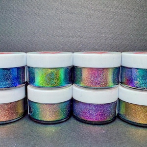 May include: Eight small, clear jars filled with iridescent glitter. The glitter shimmers with a rainbow of colors, including blue, green, purple, and gold. Each jar has a white lid. The jars are arranged in two rows of four.