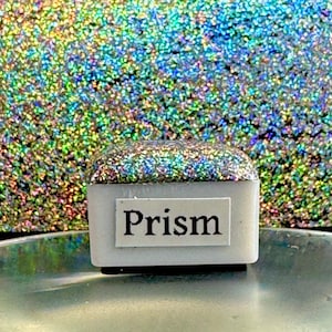 以下が含まれることがあります： 「Prism」の文字が印刷された、小さな白い長方形の物体。物体の上面は虹色のグリッターで覆われています。背景は、きらめくマルチカラーの表面です。