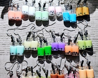 Może przedstawiać: Zestaw 18 par wiszących kolczyków z miniaturowymi kubkami boba tea z kolorowymi płynami i czarnymi perełkami tapioki. Kolczyki są wykonane z polimerowej gliny i mają srebrne haczyki.