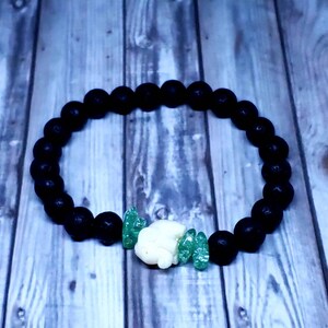 May include: Black lava rock beaded bracelet with a white and green charm. The charm is a small, carved animal.