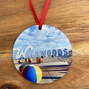 May include: Round ornament with a picture of the Wildwoods sign and beach balls. The sign is blue and white with the word "Wildwoods" in white letters. The beach balls are red, yellow, blue, and green. The background is a blue sky with white clouds.