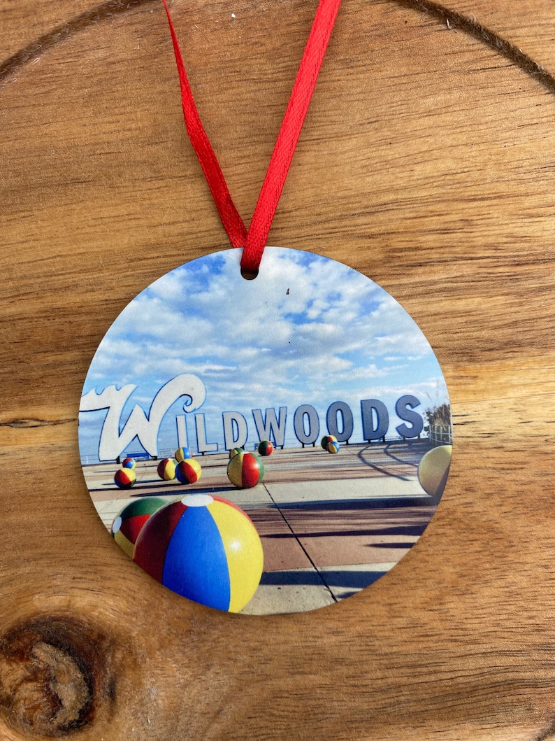 May include: Round ornament with a picture of the Wildwoods sign and beach balls. The sign is blue and white with the word "Wildwoods" in white letters. The beach balls are red, yellow, blue, and green. The background is a blue sky with white clouds.