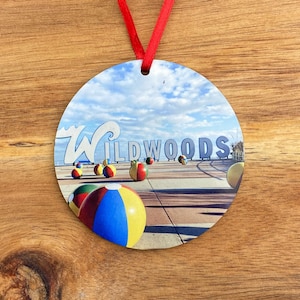 May include: Round ornament with a photo of the Wildwoods sign and beach balls on the boardwalk. The sign is blue and white with the word "Wildwoods" in white letters. The beach balls are red, yellow, blue, and green.