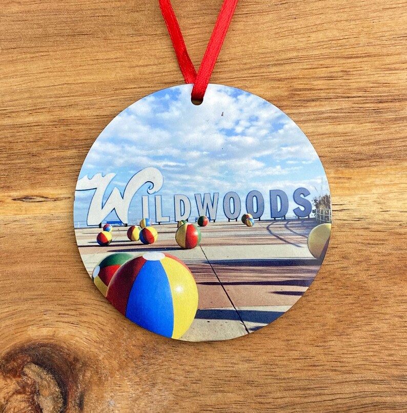 May include: Round ornament with a photo of the Wildwoods sign and beach balls on the boardwalk. The sign is blue and white with the word "Wildwoods" in white letters. The beach balls are red, yellow, blue, and green.
