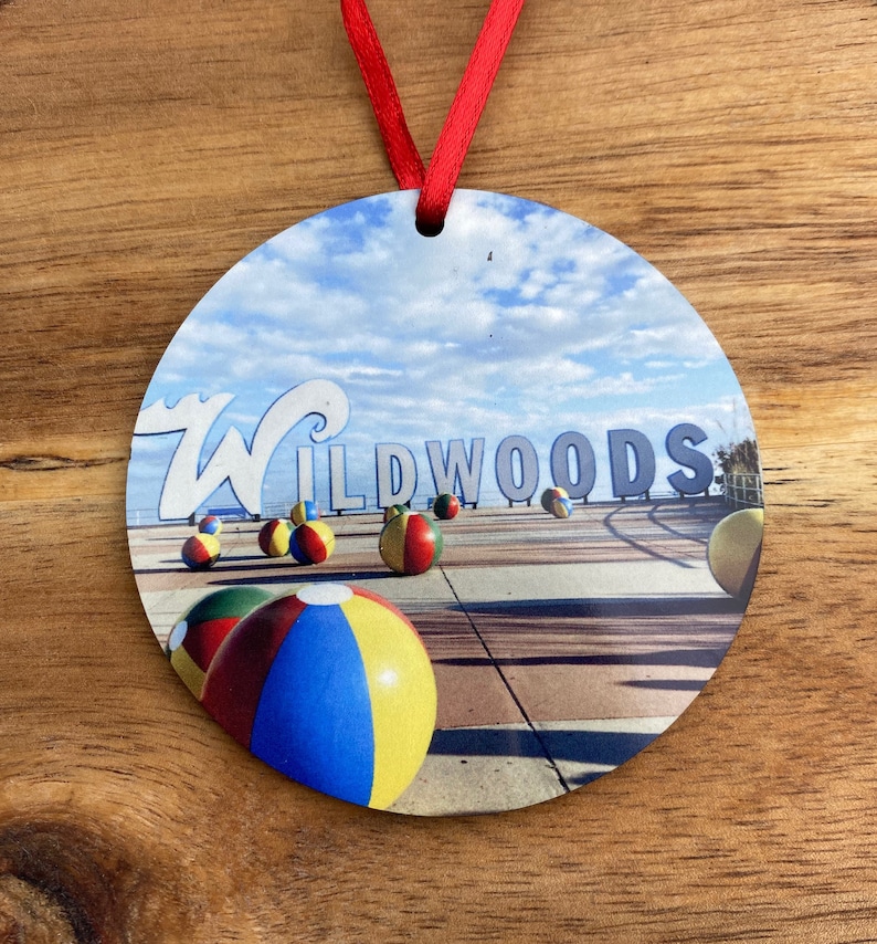 May include: Round ornament with a picture of the Wildwoods sign and beach balls. The sign is white with blue and green lettering. The beach balls are red, yellow, blue, and green.