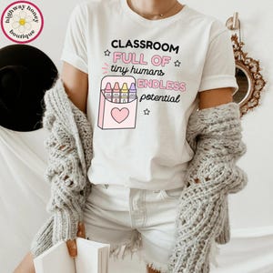 May include: A white short-sleeved t-shirt featuring the black and pink text CLASSROOM FULL OF tiny humans ENDLESS potential and a graphic of a pink crayon box containing four colorful crayons with a heart icon and small star accents.