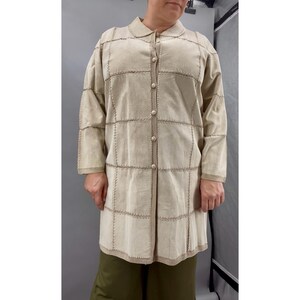 May include: A beige, long-sleeved coat with a collared neckline and button closure. The coat features a patchwork design with visible stitching and a textured appearance. The coat's length extends to the mid-thigh.